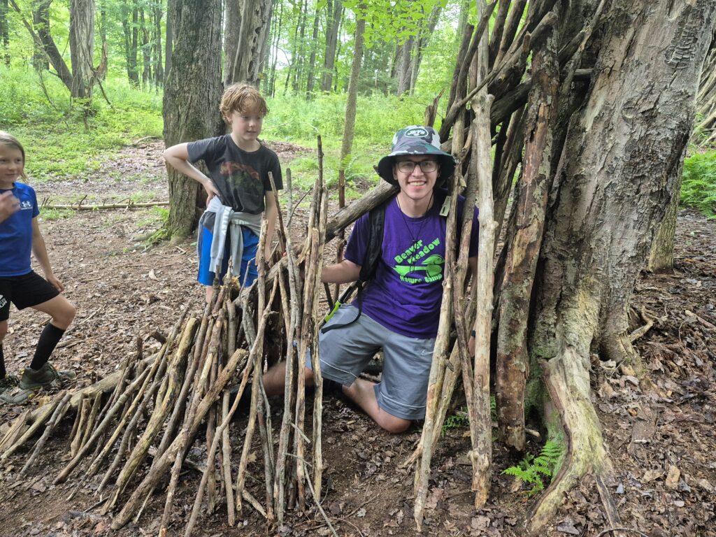 A camp counselor smiling in a stick shelter, as two other campers stand nearby